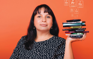 Professor Cecilia Martinez-Gomez behind a peach background, she is holding up a stack of cellphones in one hand as digital graphics of elements displayed on the periodic table arise from the phones.