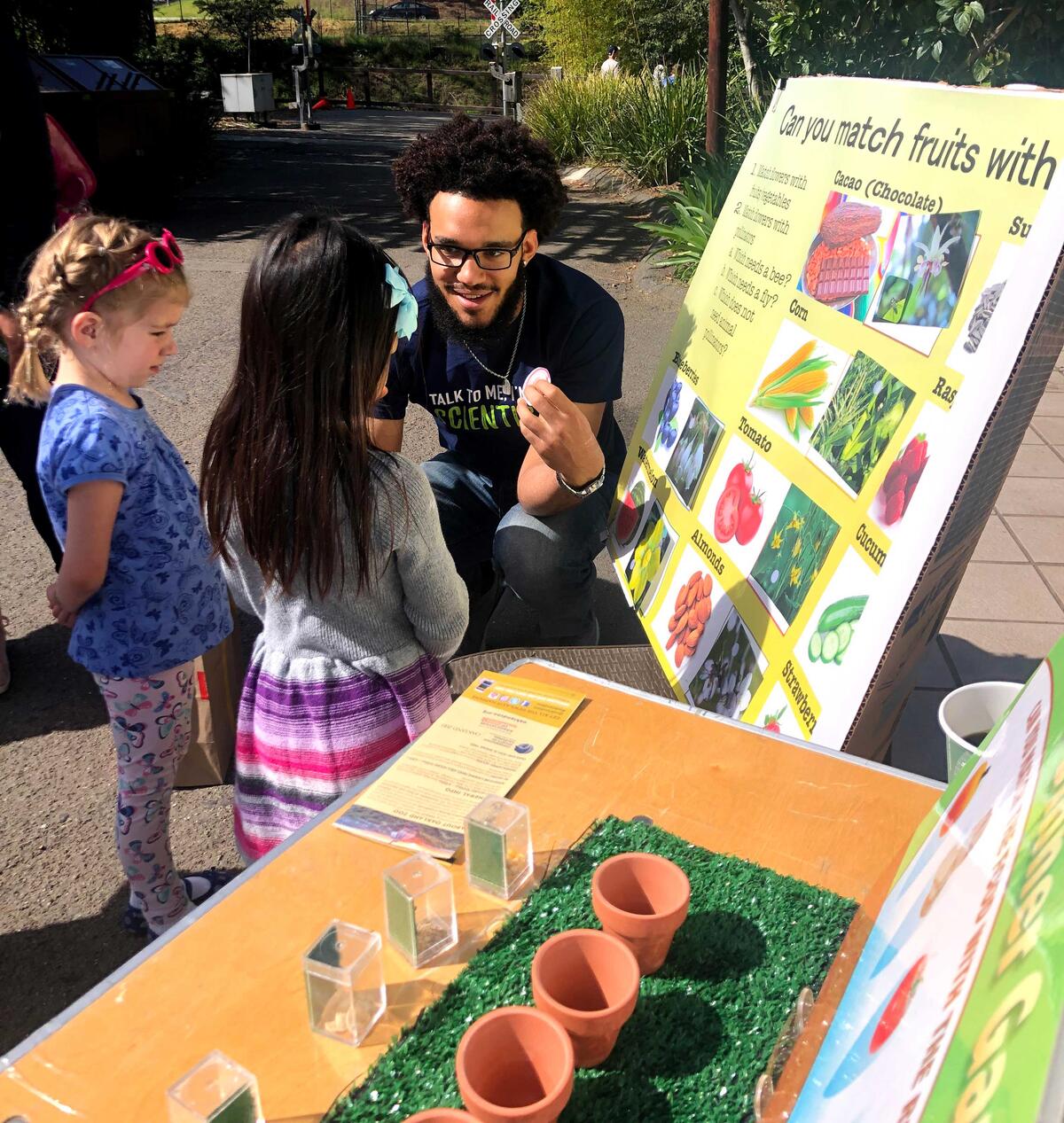 Man crouches down to teach two small children about fruits and vegetables, poster in background
