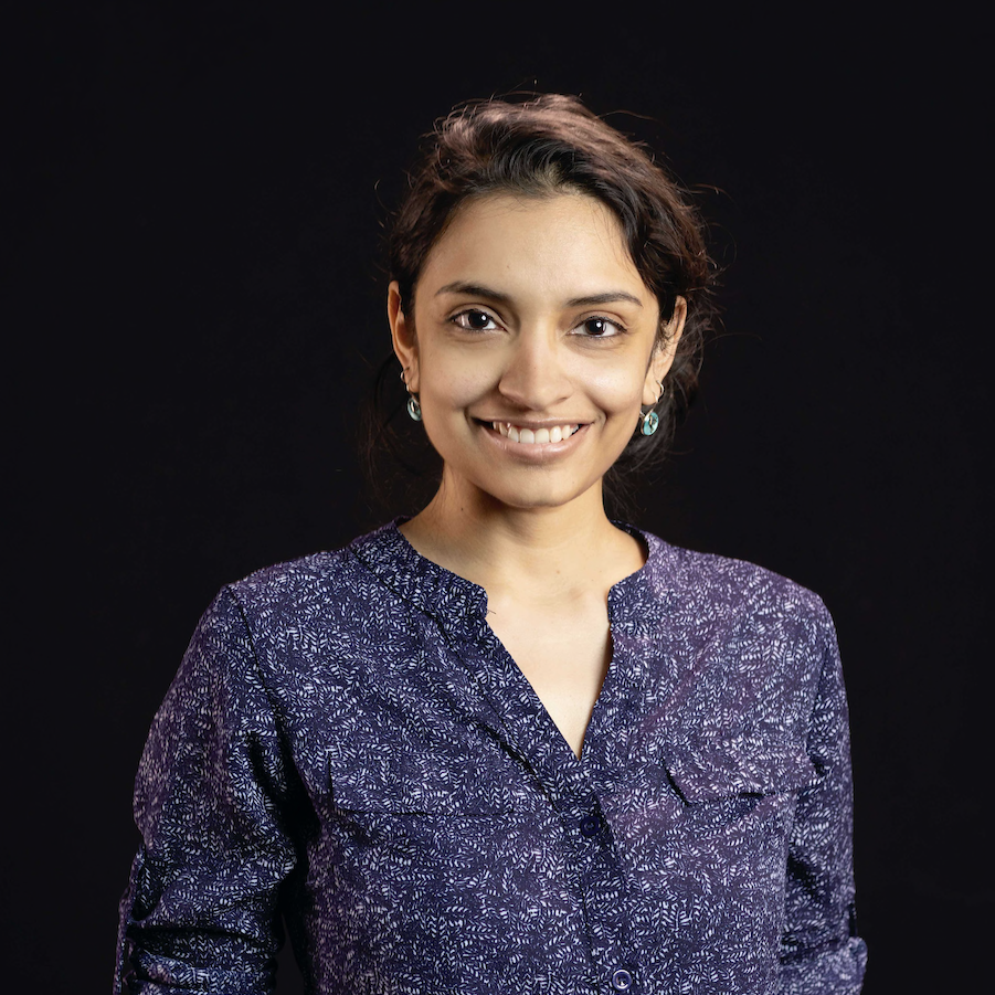 Smiling South Asian woman with black hair in a bun. She is wearing a blue shirt and earrings.